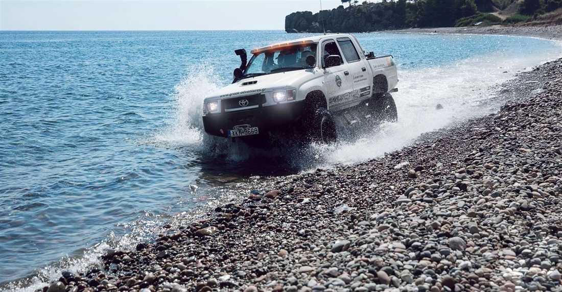 A white 4x4 pickup splashing through shallow sea water along a pebble beach in Halkidiki.