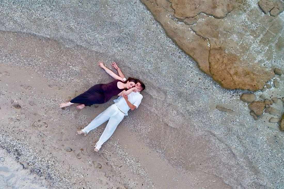 A couple lying on a pebble beach in white and dark clothes, photographed from above by a drone beside rocks and clear water