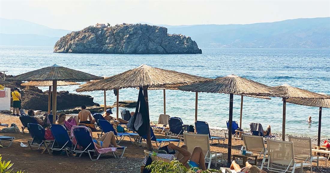 Straw umbrellas and sunbeds on a pebble beach on Hydra, facing calm blue sea and a rocky islet.