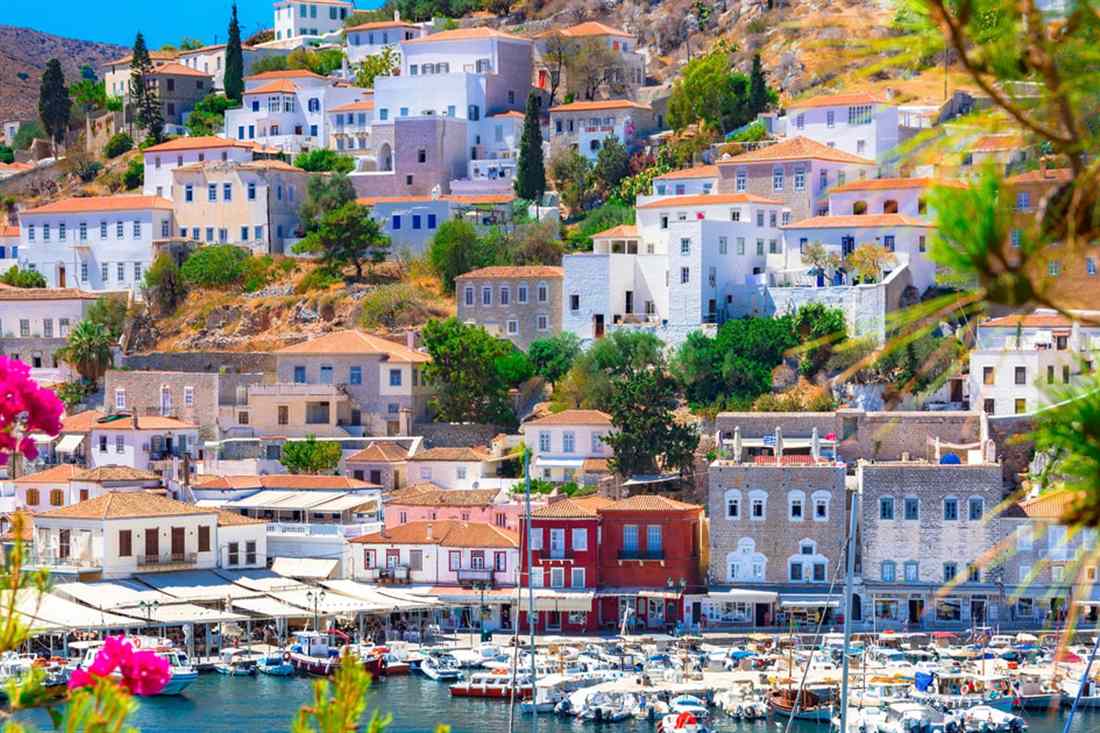 Hydra town rising in tiers of stone mansions and white houses above the harbour, with moored boats and pink flowers in the foreground.