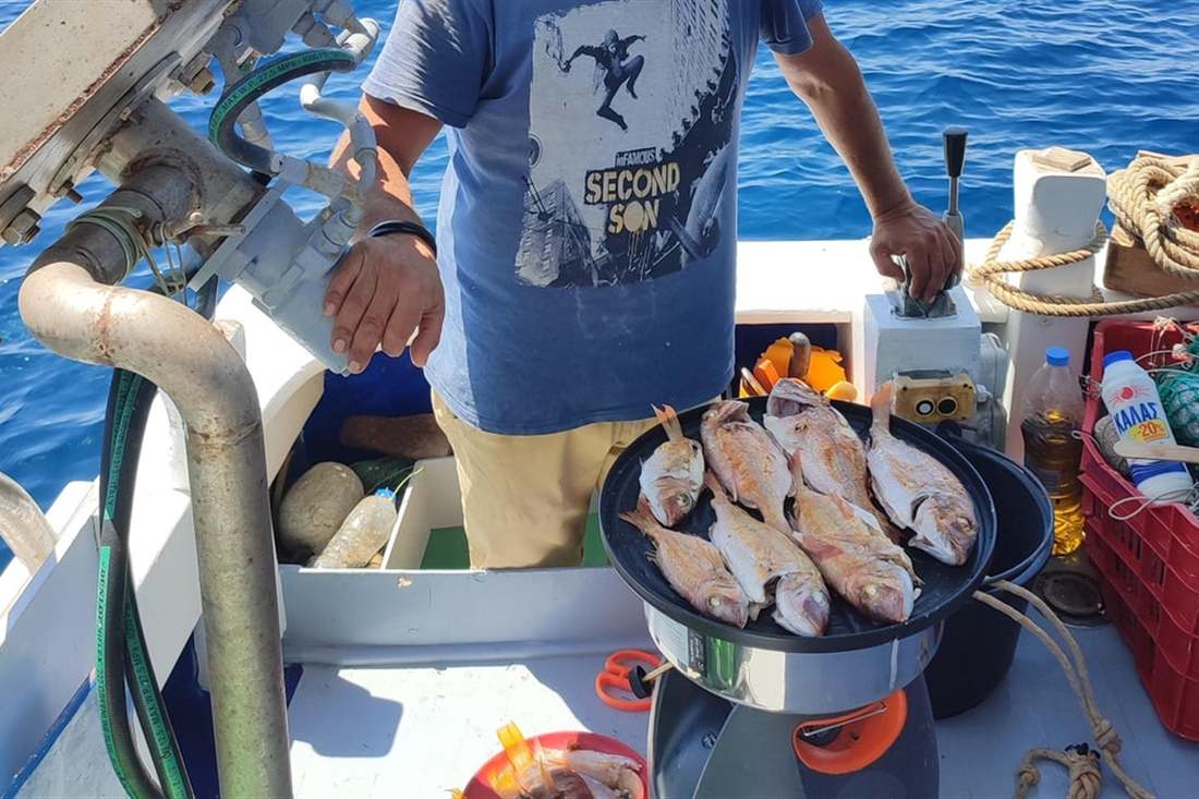 A man fries a pan of red fish on a gas ring on the deck of a fishing boat, with the blue sea behind, on a Hydra fishing trip.