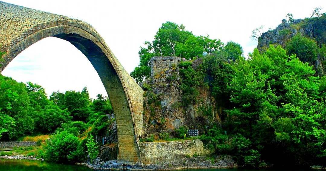 The tall single-arch stone bridge of Konitsa over the green river, with a rock and trees behind.