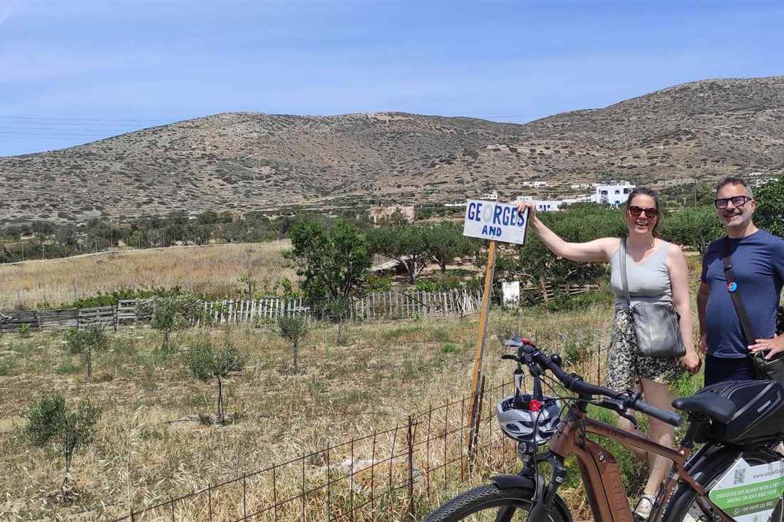 A couple beside a hand-painted sign and an e-bike in the olive groves of Pano Kampos on Ios