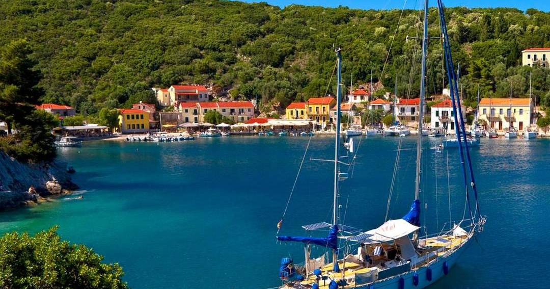 A sailing yacht moored in a green bay in front of a harbour village with red-roofed houses on Ithaca.