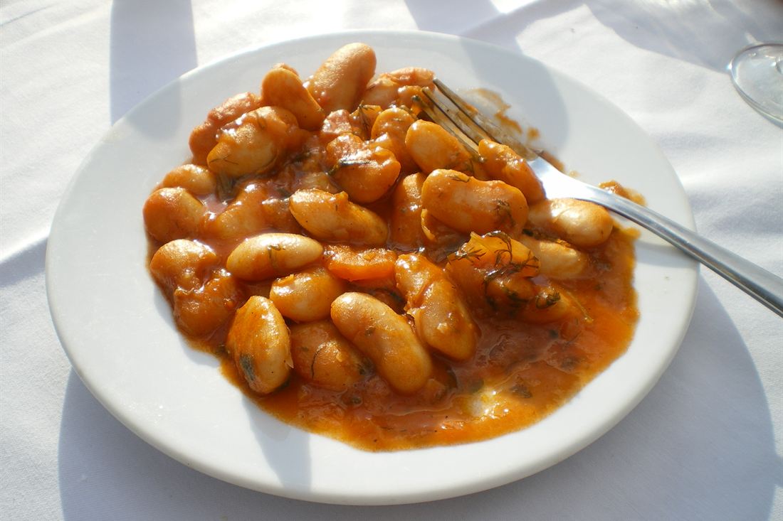 White plate of gigandes plaki, giant white beans baked in tomato sauce with dill, on a white tablecloth in bright sun