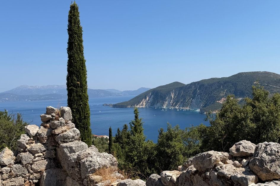 Tall cypress and old stone walls above a deep blue bay with green hills and distant mountains on Ithaca