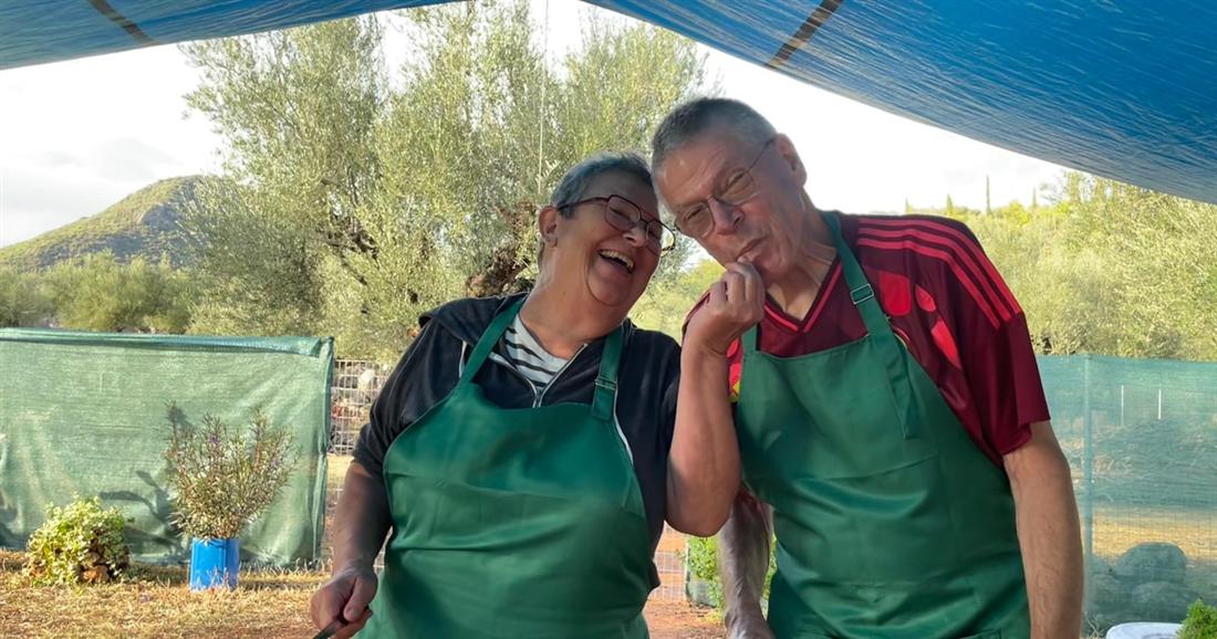 Two cooks in green aprons at the open-air kitchen of a Kalamata cooking class in an olive grove.
