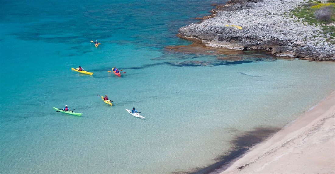 Sea kayakers paddling yellow, red and green kayaks over turquoise water off a white beach on the Mani coast