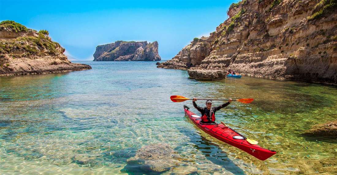 A paddler raising the paddle in a red sea kayak in a turquoise rocky cove of Navarino Bay, with a rock islet behind