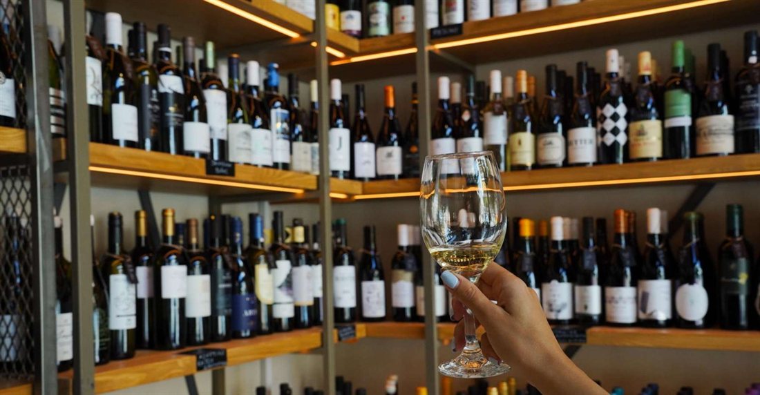 A glass of white wine raised in front of lit wooden shelves full of wine bottles