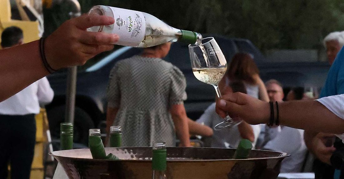 A hand pours chilled white wine from a bottle into a tasting glass above an ice bucket of wine bottles at an outdoor tasting