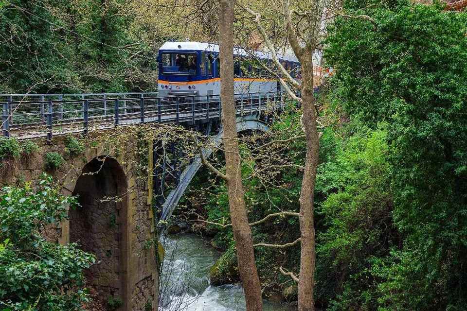 A blue and white Odontotos rack railway train crossing a stone bridge over the river in the green Vouraikos gorge.