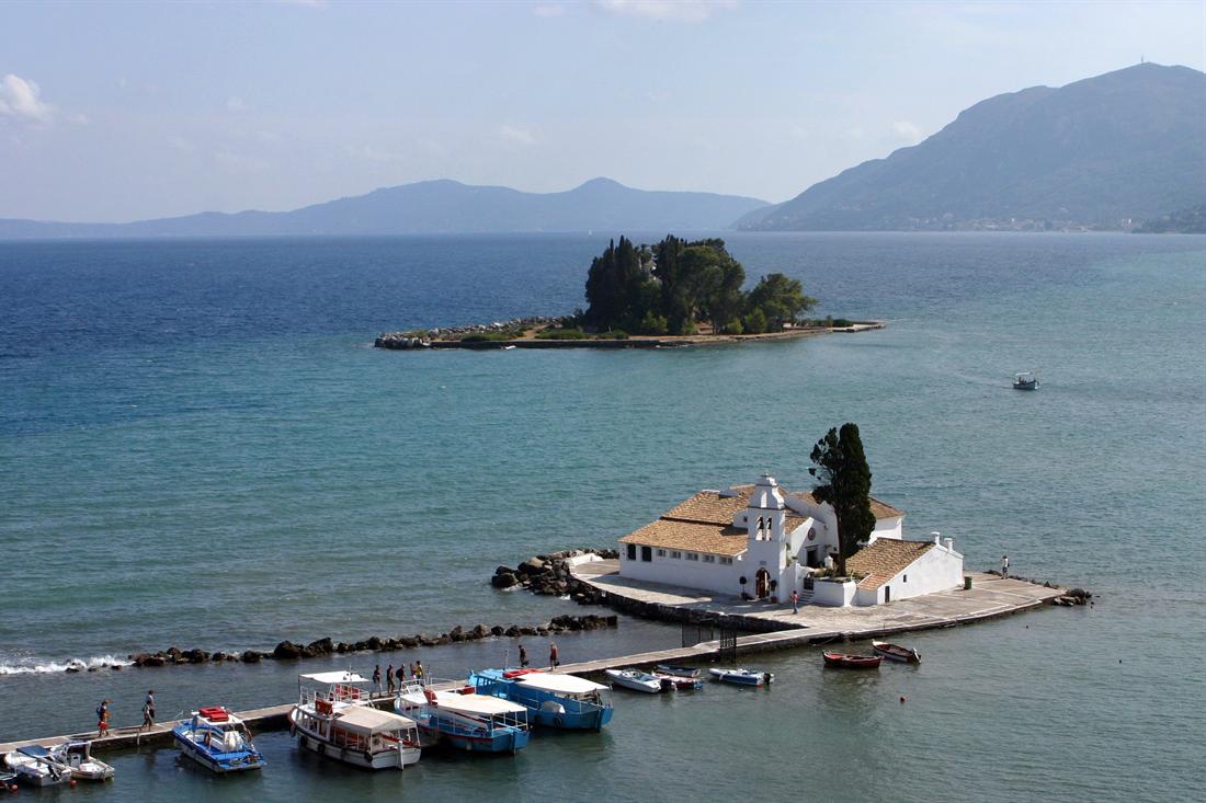 Vlacherna Monastery on its causeway islet and the wooded Mouse Island in a blue bay, with tour boats moored along the causeway and mountains behind