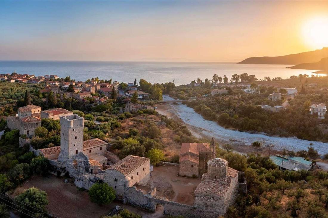 Old stone tower and churches above a dry riverbed beside a seaside village and bay at sunset on the Messenian Mani coast