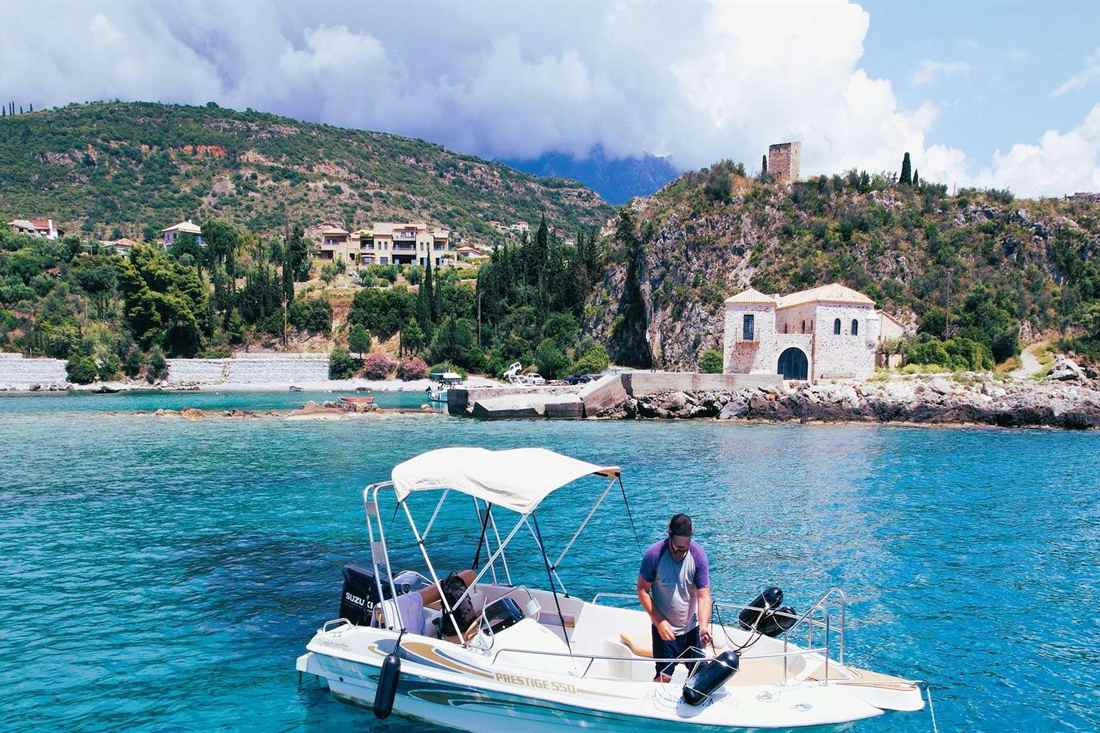 A white rental motorboat on clear blue water off Kardamyli, with a stone tower and a stone church on the shore behind