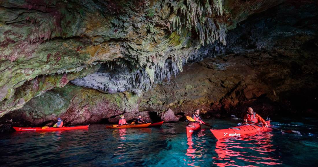 Paddlers in red sea kayaks inside a sea cave with clear blue water, on a Kardamyli sea kayaking tour.