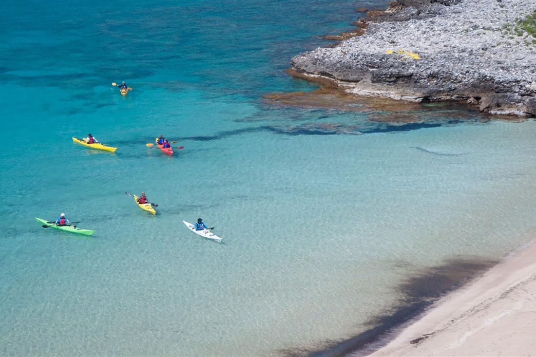 Sea kayakers paddling across a shallow turquoise cove beside a pale rocky shore on Kardamyli sea kayaking.