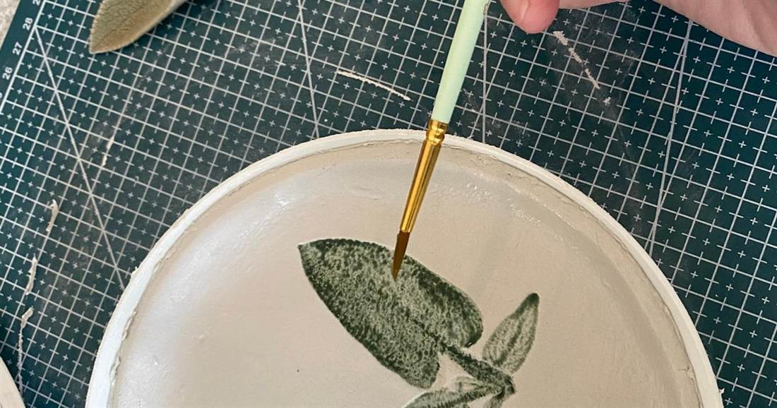 A brush adding green glaze to a leaf print on a clay plate in a Karpathos pottery class.