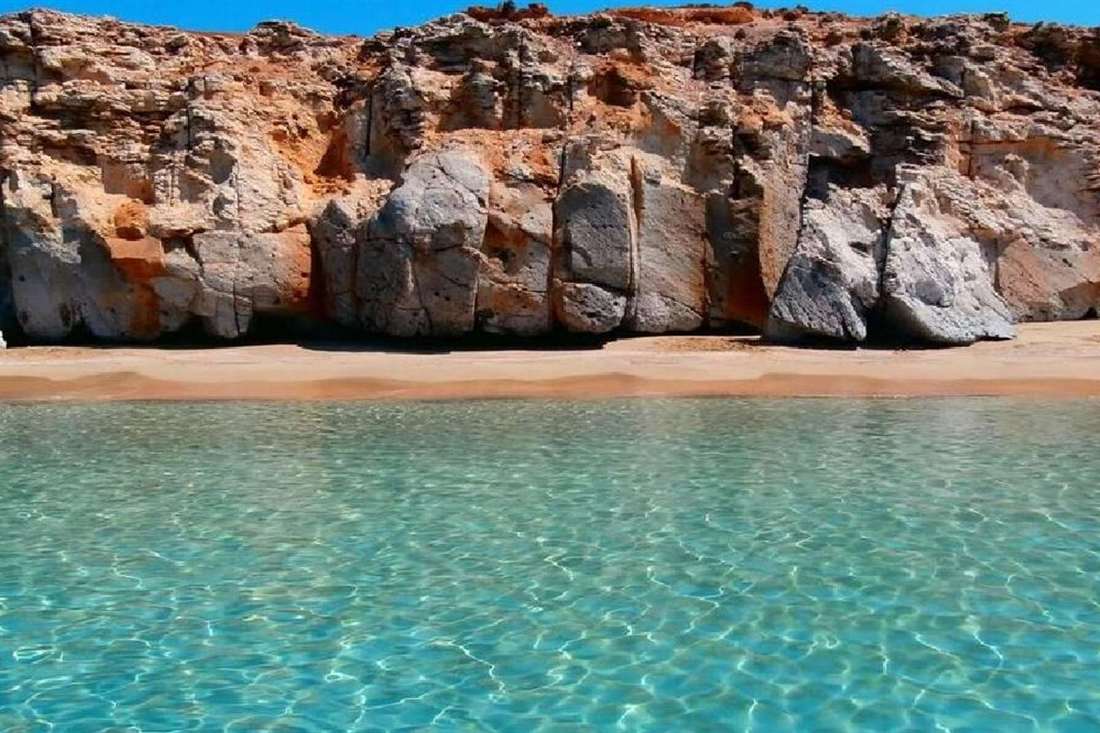 Orange and grey sea cliffs above a sandy cove and clear turquoise water