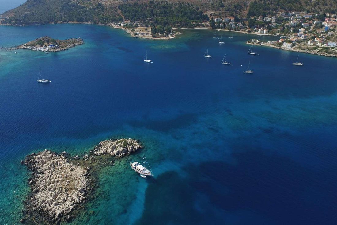 Aerial view of a white tour boat anchored beside a rocky islet in the blue bay of Kastellorizo, with sailing yachts moored off the harbour town of the island