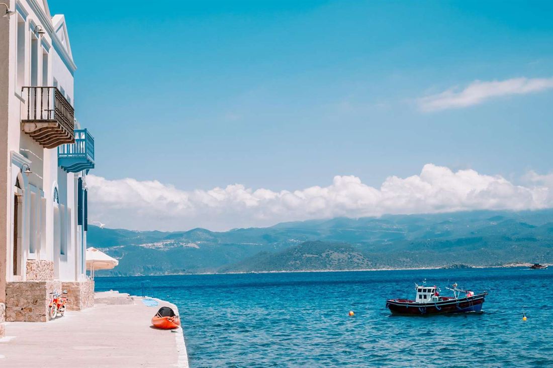 Small red and navy boat on the sea beside a white quay and balconied house on Kastellorizo, with mountains on the far shore under a blue sky
