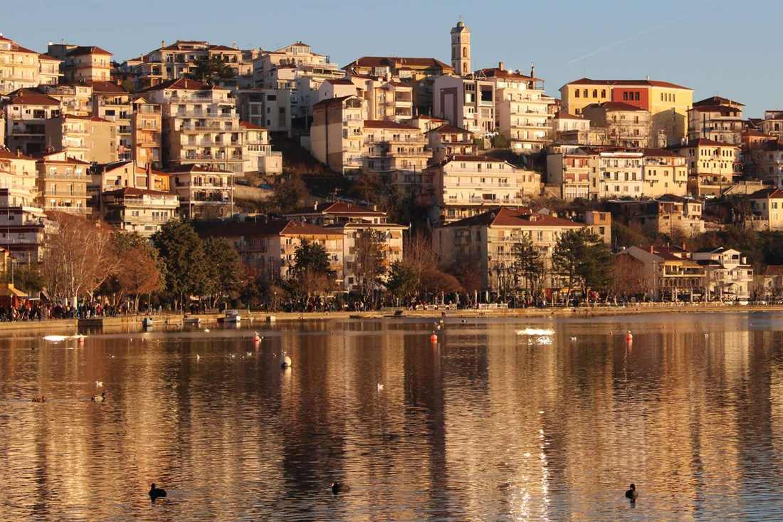 Hillside houses and a church bell tower in Kastoria lit by low sun above Lake Orestiada, with the town reflected in the water and dark ducks floating on the lake