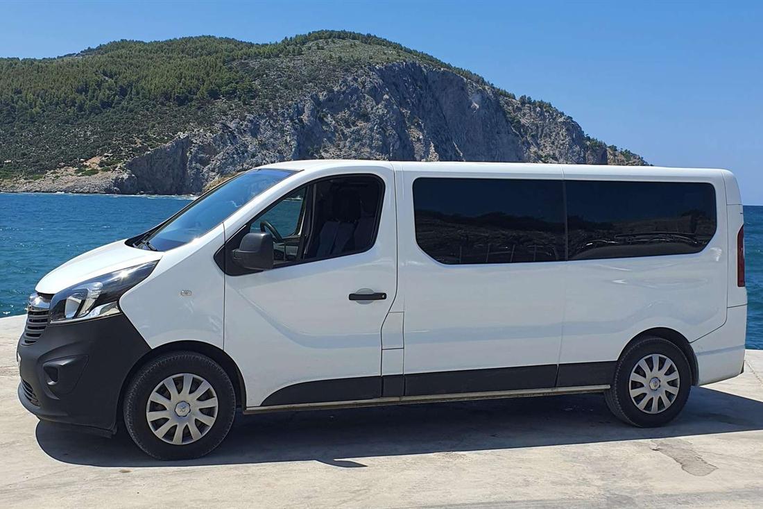 White passenger van parked on a sunny seafront quay with blue sea and a rocky green headland behind
