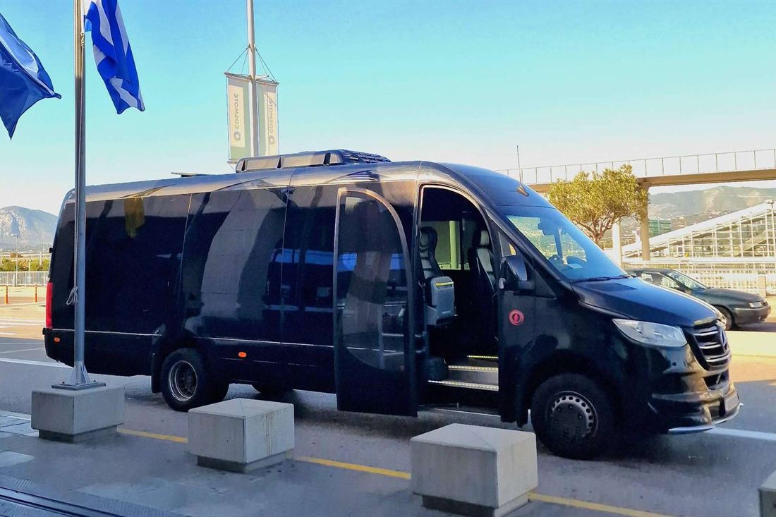 Black minibus with its side door open at an airport kerb beside two Greek flags
