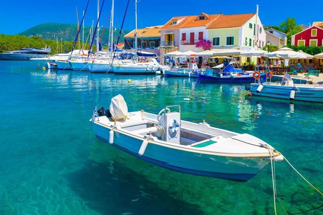 The harbour of Fiskardo on Kefalonia, with pastel houses, cafe umbrellas, moored sailing boats and a small motorboat in clear water.