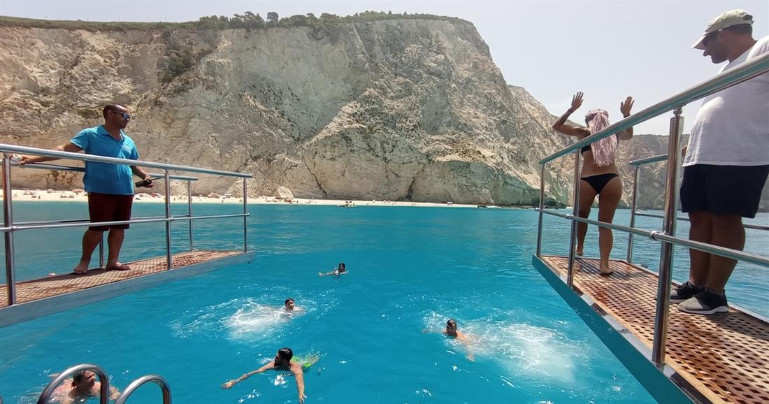 Swimmers jump from a cruise boat into clear blue water below white cliffs in the Ionian Sea.
