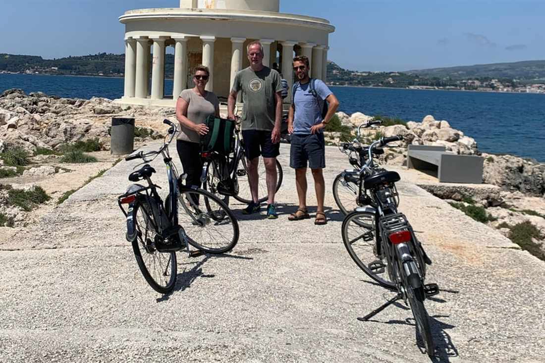 Three cyclists stand beside their bikes on the paved path to the round, columned lighthouse at Argostoli, on a Kefalonia e-bike tour.