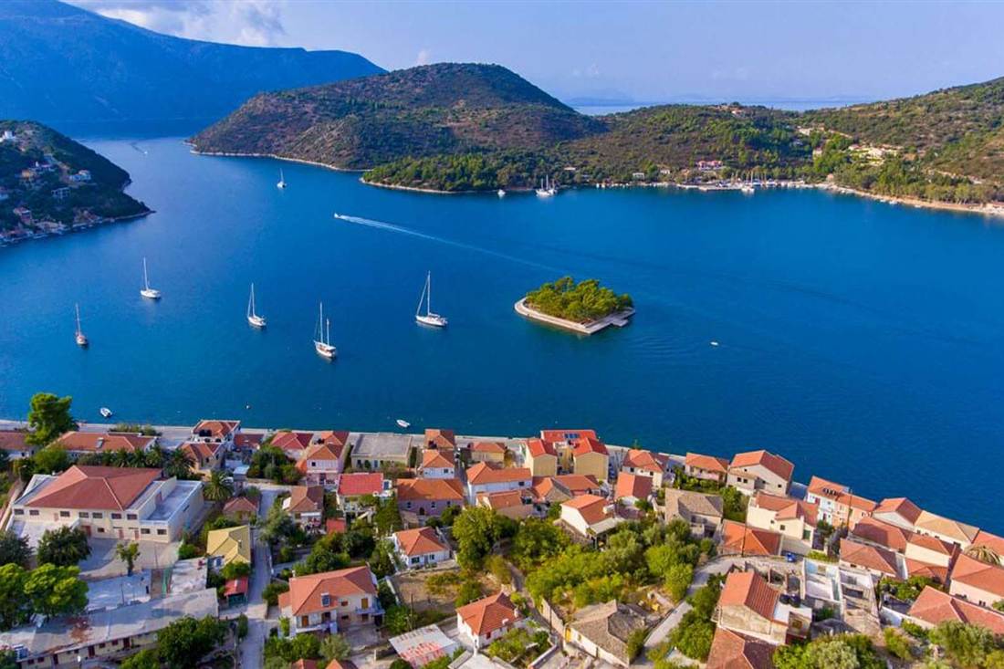 Aerial view of a blue bay with sailing boats, a small wooded islet and red-roofed houses on a hillside shore