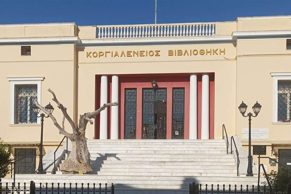 Cream-coloured library building with a red entrance, white columns, marble steps and an olive-tree sculpture in front