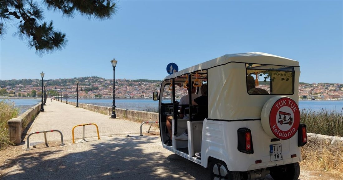 A tuk tuk on a long stone bridge with lamp posts over the water, with a town on the far shore, on a Kefalonia tuk tuk tour.