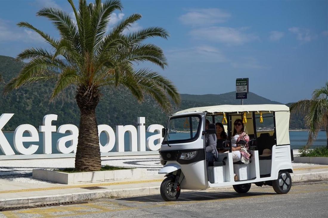 A tuk tuk with passengers beside a large Kefalonia letter sign and a palm tree on a Kefalonia tuk tuk tour.