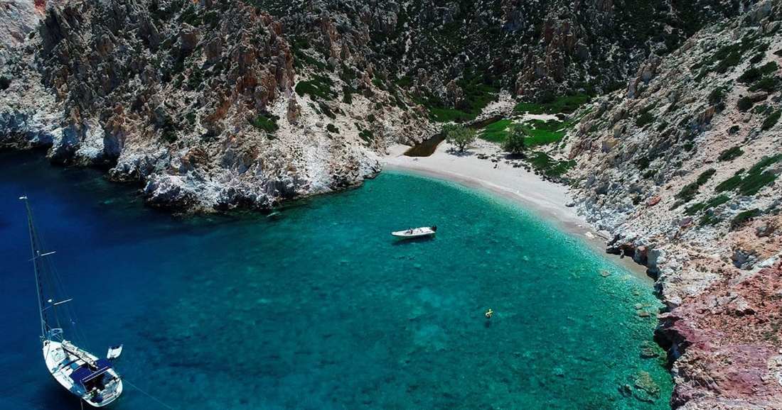 A sailing boat and a small motorboat anchored in a green-blue cove below rocky cliffs, beside a small sand beach.