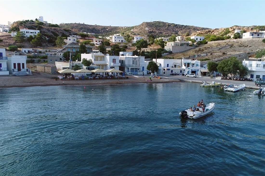 A small inflatable boat with passengers heads for the waterfront of a white-house harbour village on Kimolos, with tavernas on the beach.