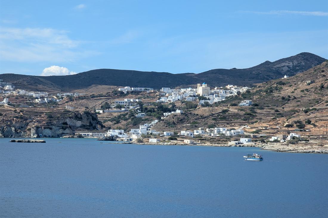 White houses and a domed church on terraced hills above a calm blue bay on Kimolos, with a small blue and white boat on the water