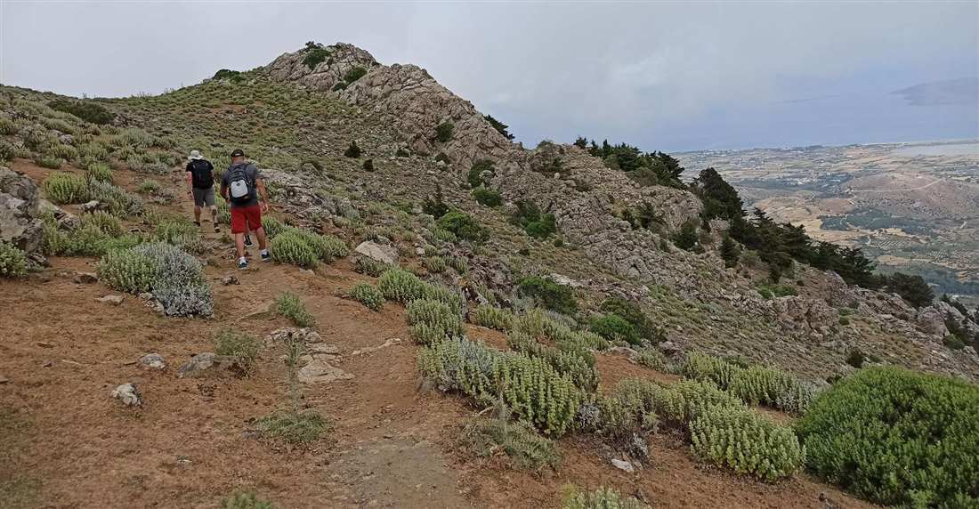 Two hikers climbing a rocky ridge path on Mount Dikaios on Kos, with the north coast and the sea below