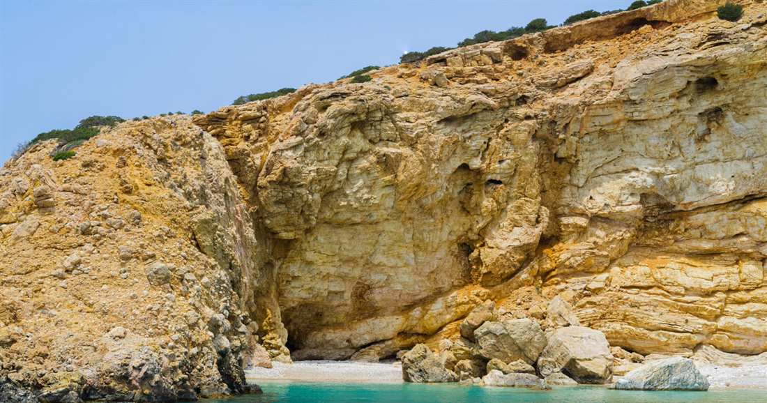 A cove of clear blue water below golden rock cliffs with a sea cave opening on Koufonisia.
