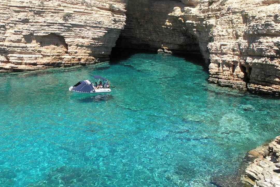A small boat in a turquoise cove below pale sandstone cliffs and a sea cave on Koufonisia.