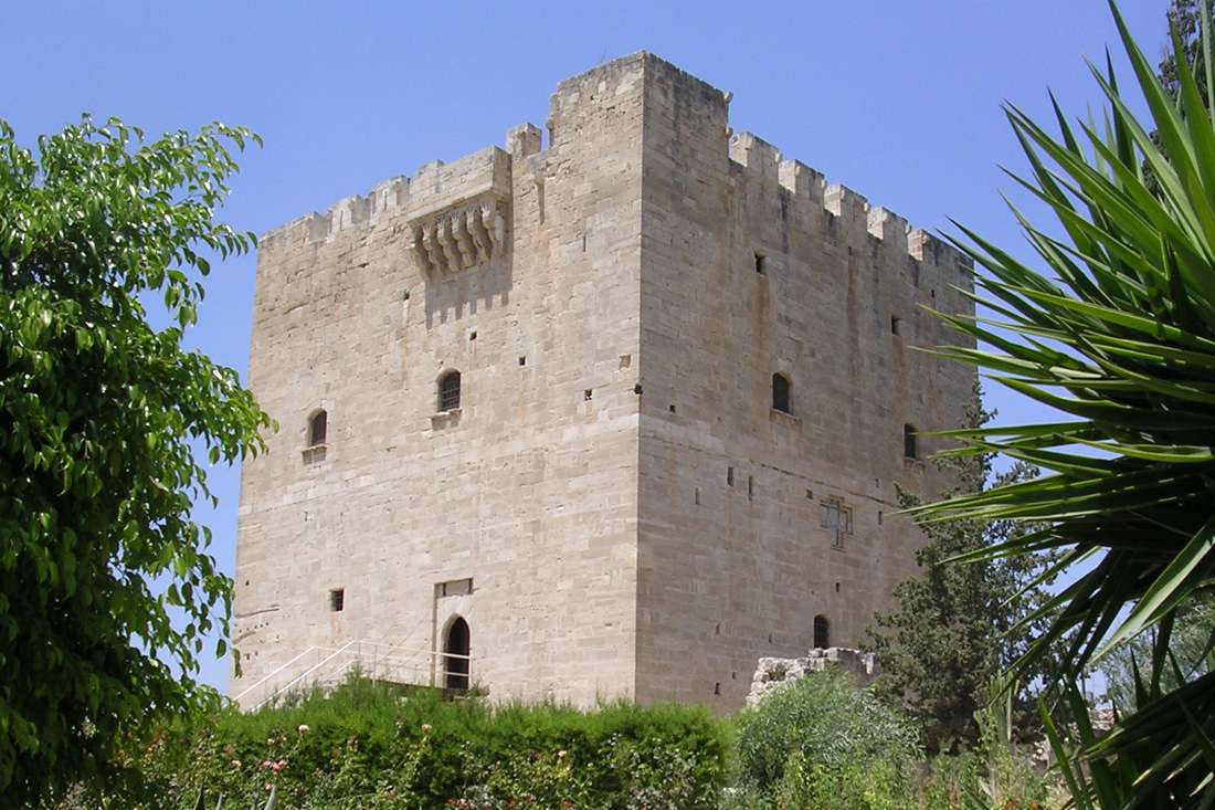 Square stone keep of Kolossi Castle with battlements and arched windows, seen through garden plants under a blue sky