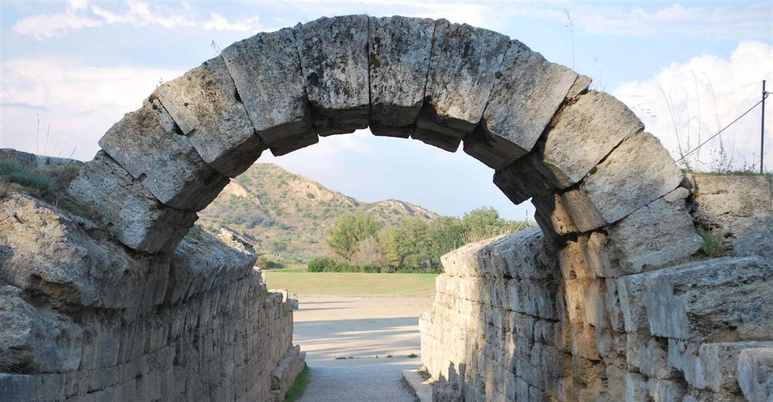 Stone arch of the vaulted tunnel leading into the ancient stadium at Olympia, with the running track and a green hill beyond