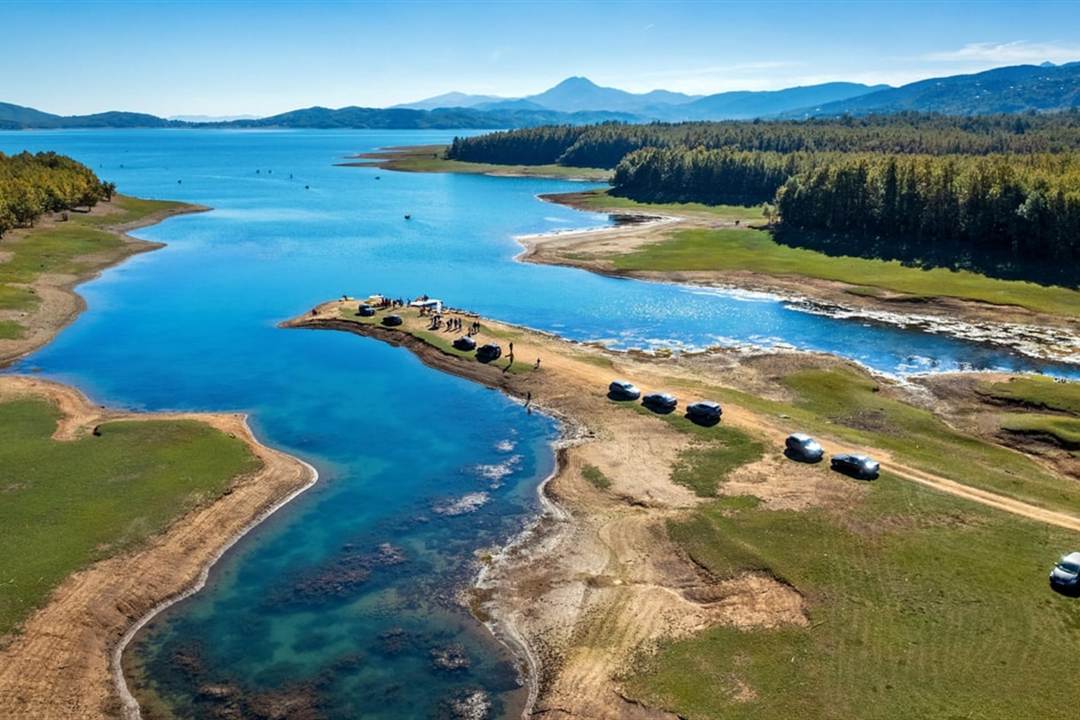 Aerial view of Lake Plastira with a winding blue inlet between green banks and fir-covered mountains behind.
