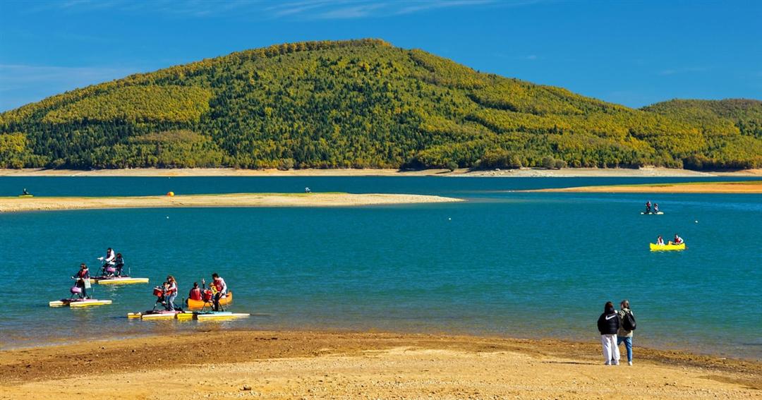 Kayaks and pedal boats on the blue water of Lake Plastira below a forested hill, with a sandy shore in front.