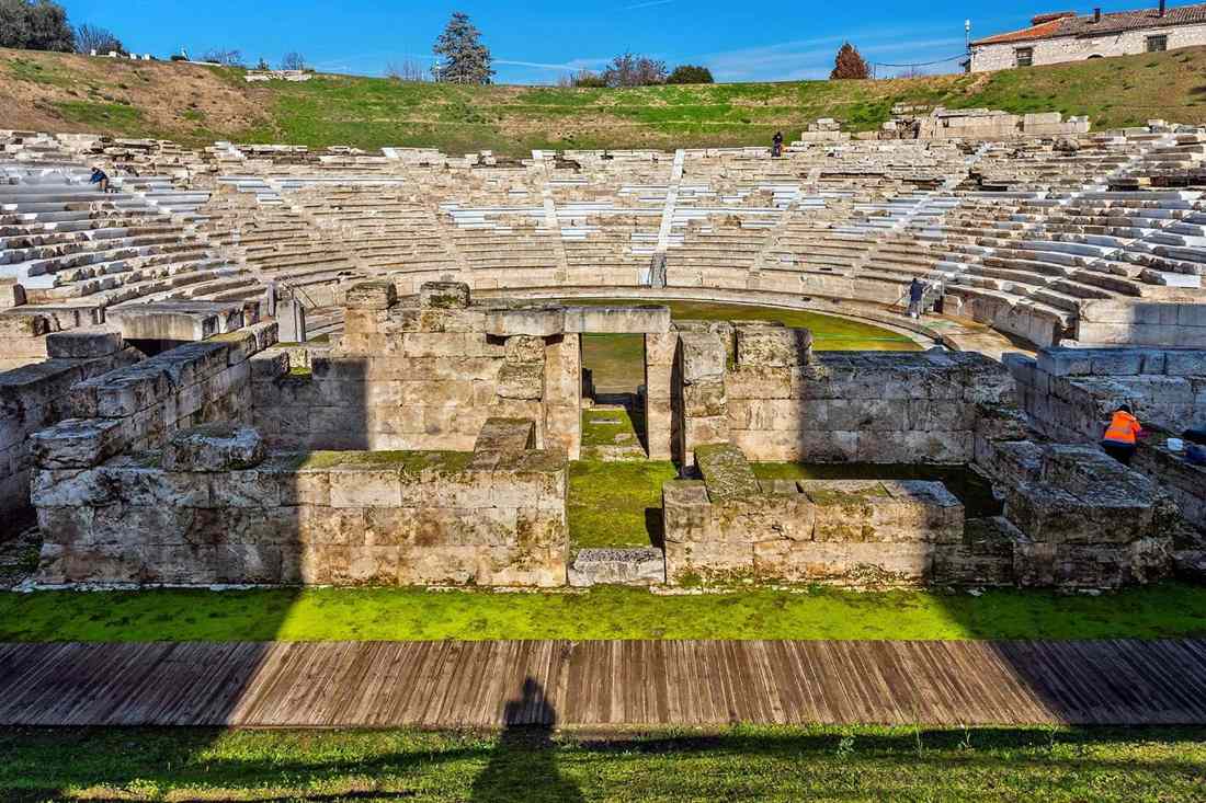 Stone stage ruins and curved rows of seats of the ancient theatre of Larissa under a blue sky