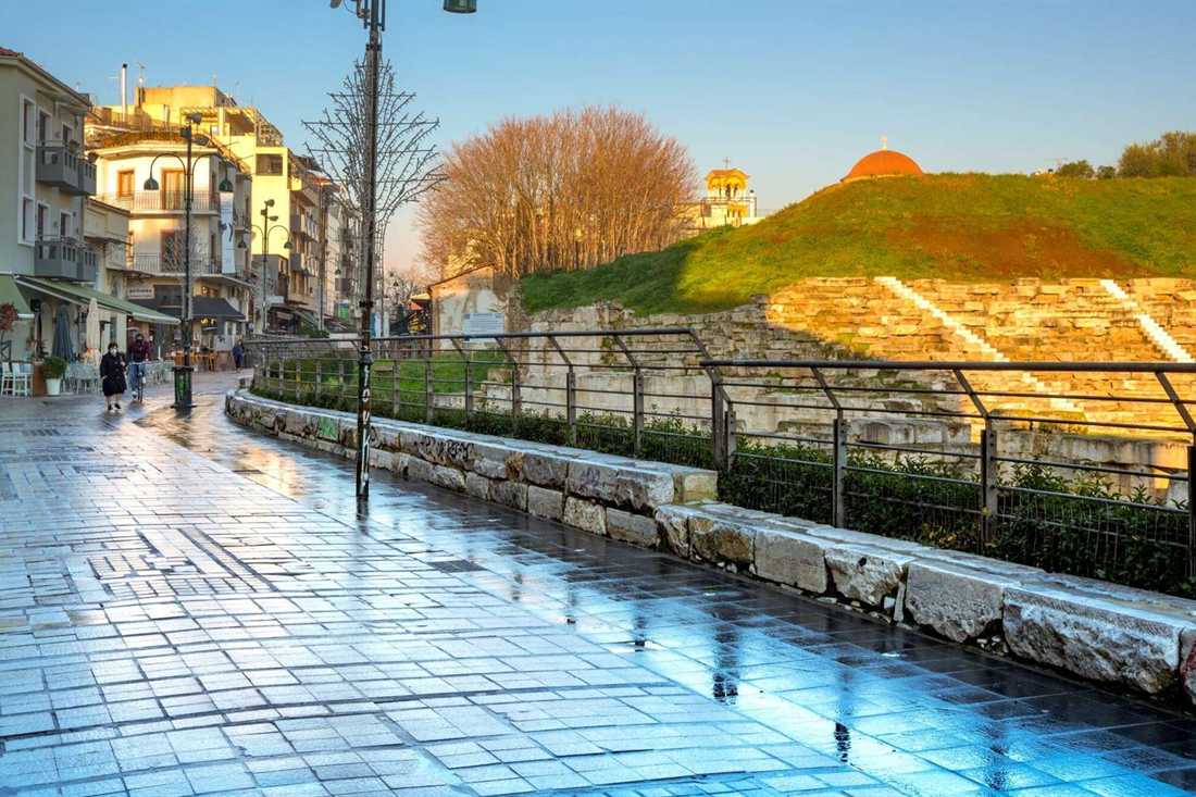 Paved pedestrian street in Larissa city centre beside the ancient theatre, with a green hill and a church dome behind it