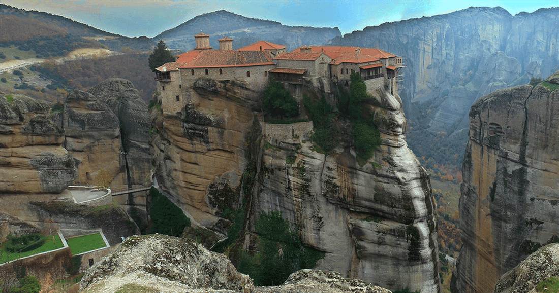 A monastery with red roofs on top of a sheer rock pillar at Meteora, with a footbridge crossing to it from the next rock.