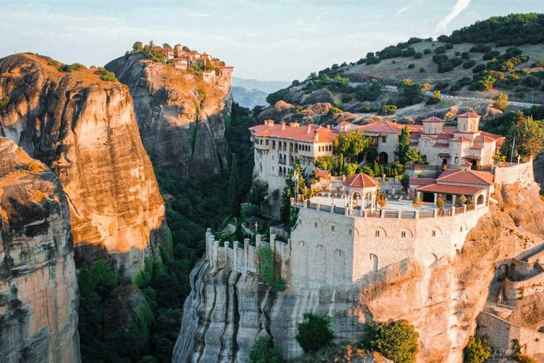 A monastery with white walls and red roofs on top of a tall sandstone rock at Meteora, beside sheer orange rock faces.