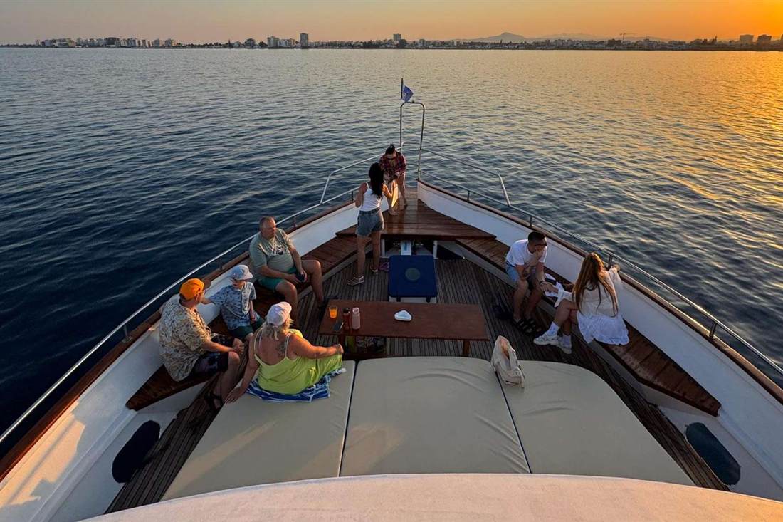 Passengers sit on the bow of a boat at sunset with the Larnaca skyline across calm water